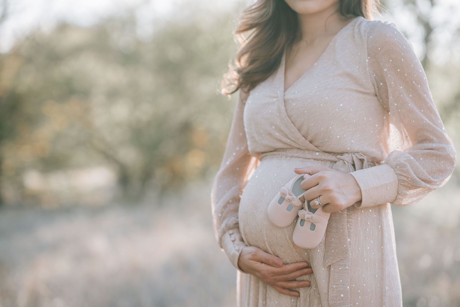 A pregnant woman in a blush wrap dress poses against a soft outdoor backdrop.
