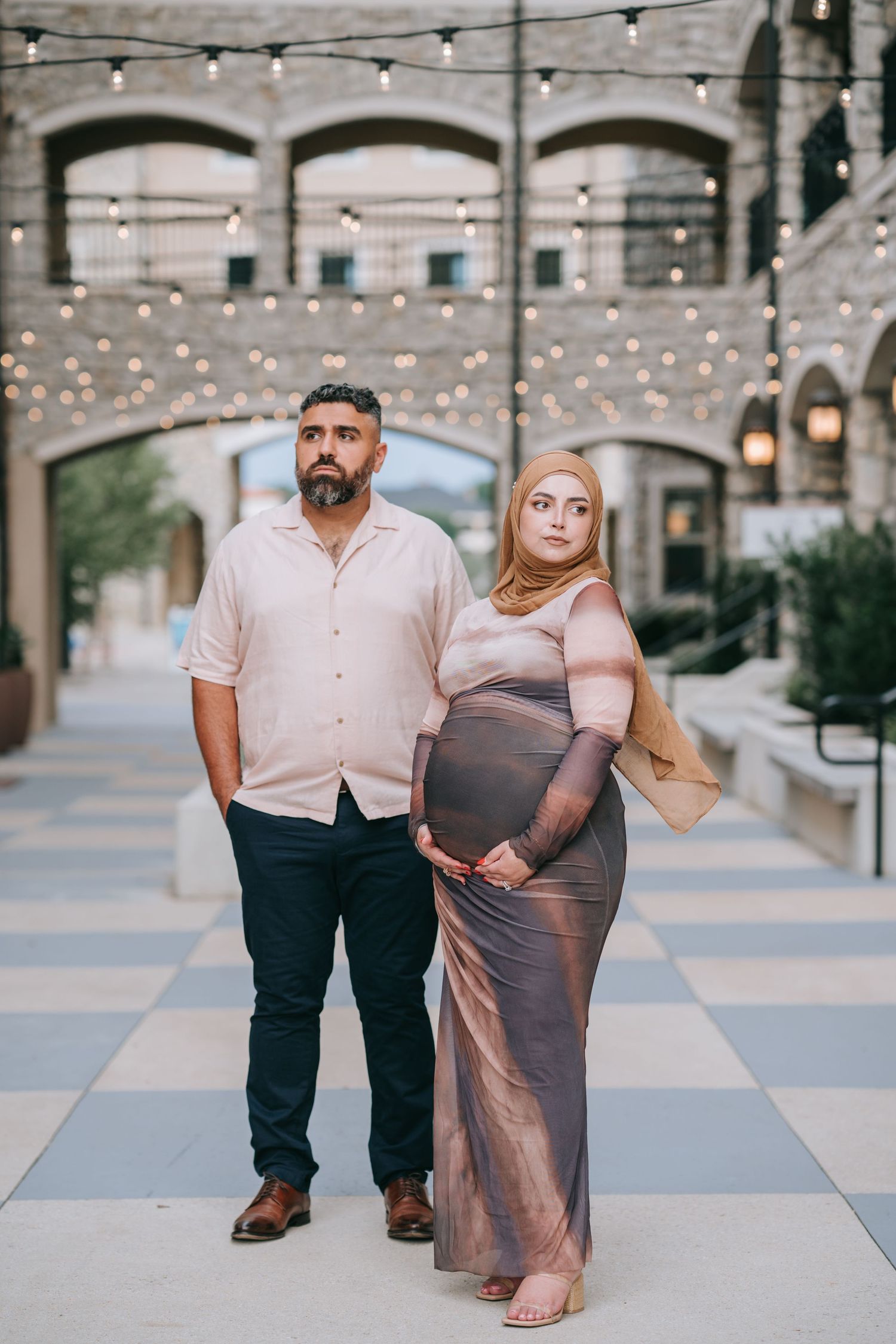 Expecting couple poses under string lights in courtyard during maternity photoshoot.