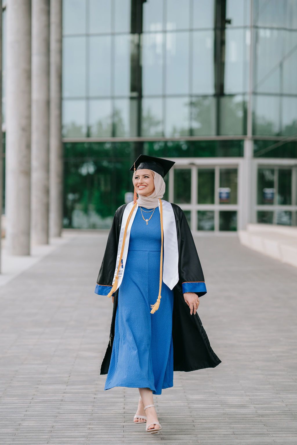 Graduate walks confidently in blue dress and black graduation gown outside modern building.