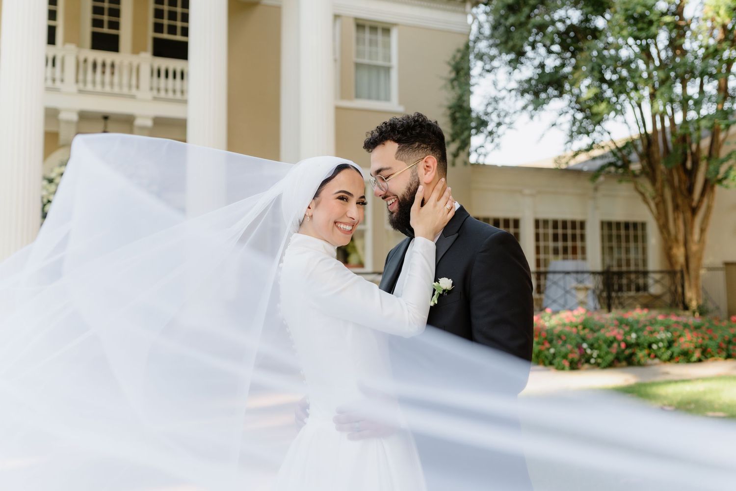 A wedding party walks together outside a white building while holding up a flowing white veil in the breeze.