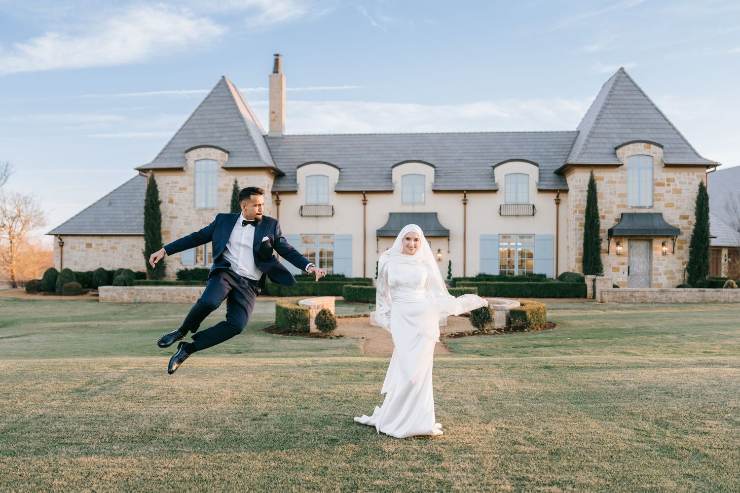 A playful moment captured on a lawn featuring a couple in wedding attire in front of a large estate mansion.