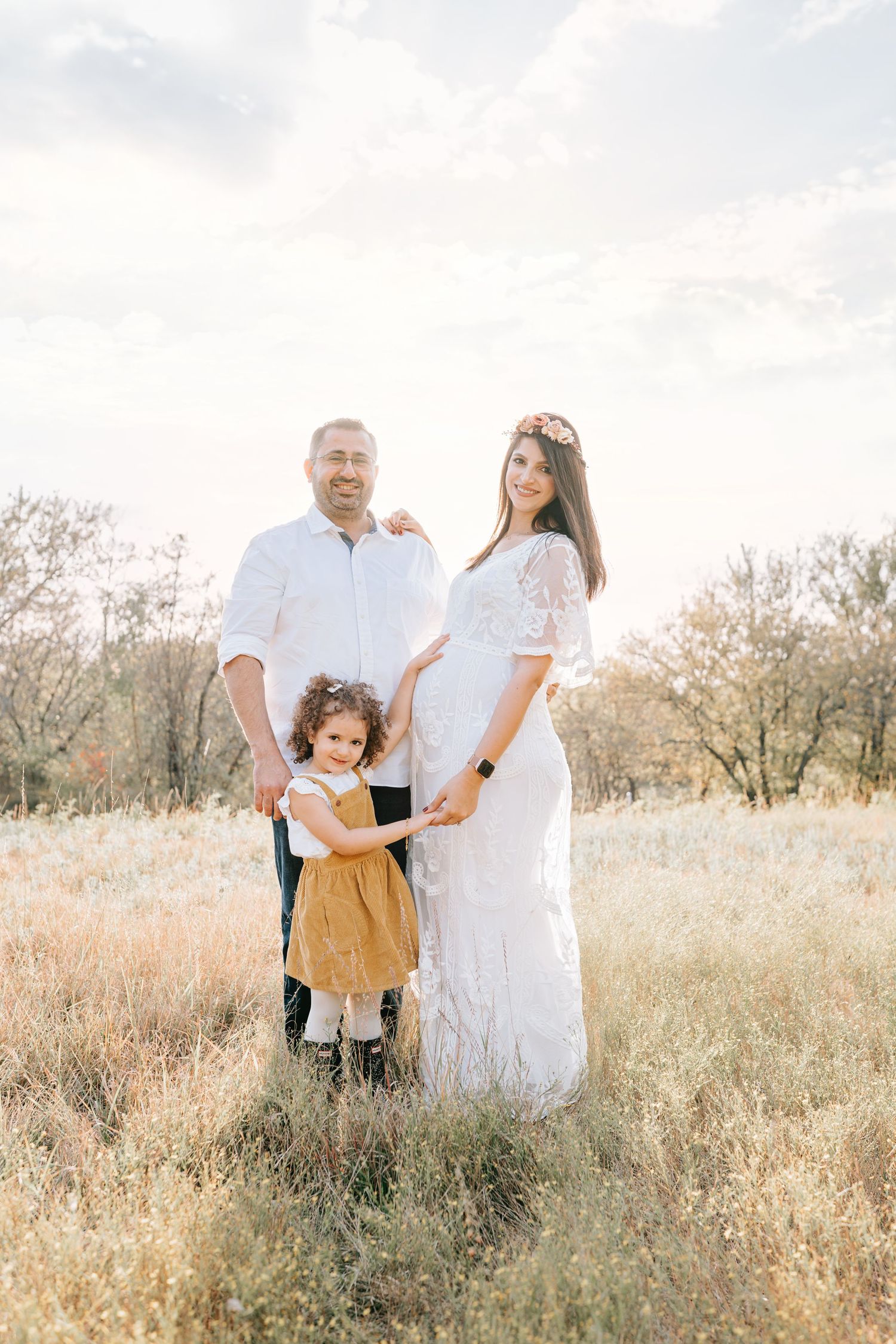 A family poses together in a golden field during sunset with a young child between them.