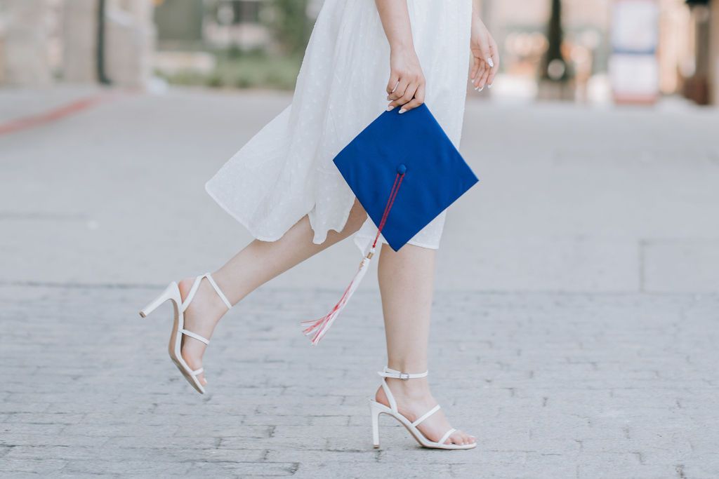 Close-up of graduate's feet in white heels holding blue diploma cover on stone pathway.