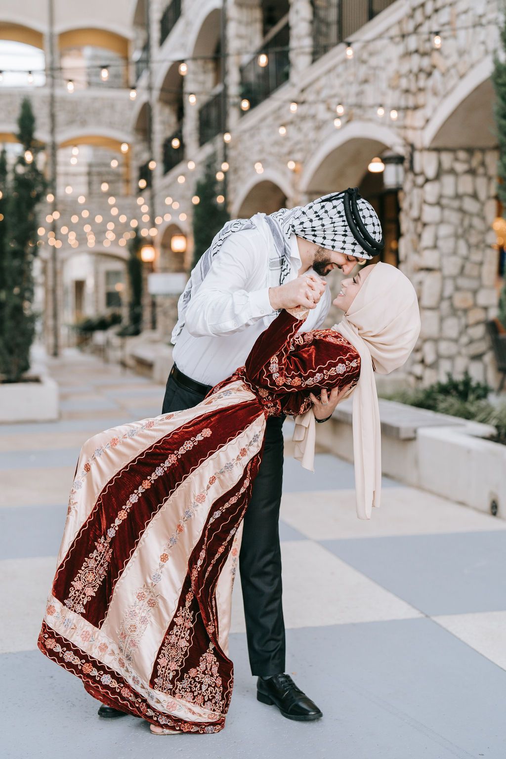 Romantic dip pose in traditional clothing under arched stone courtyard with string lights.