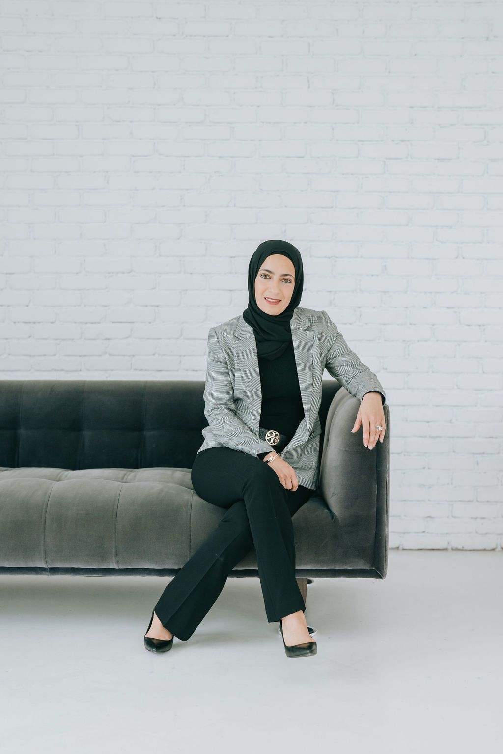 Professional wearing a grey blazer and black hijab sits elegantly on a dark leather couch against white brick wall.