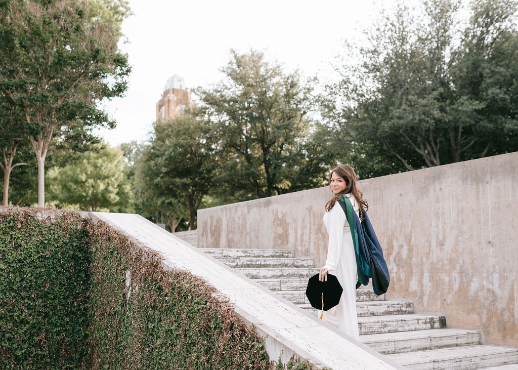 Graduate in white dress walks up stone steps with graduation cap and gown.