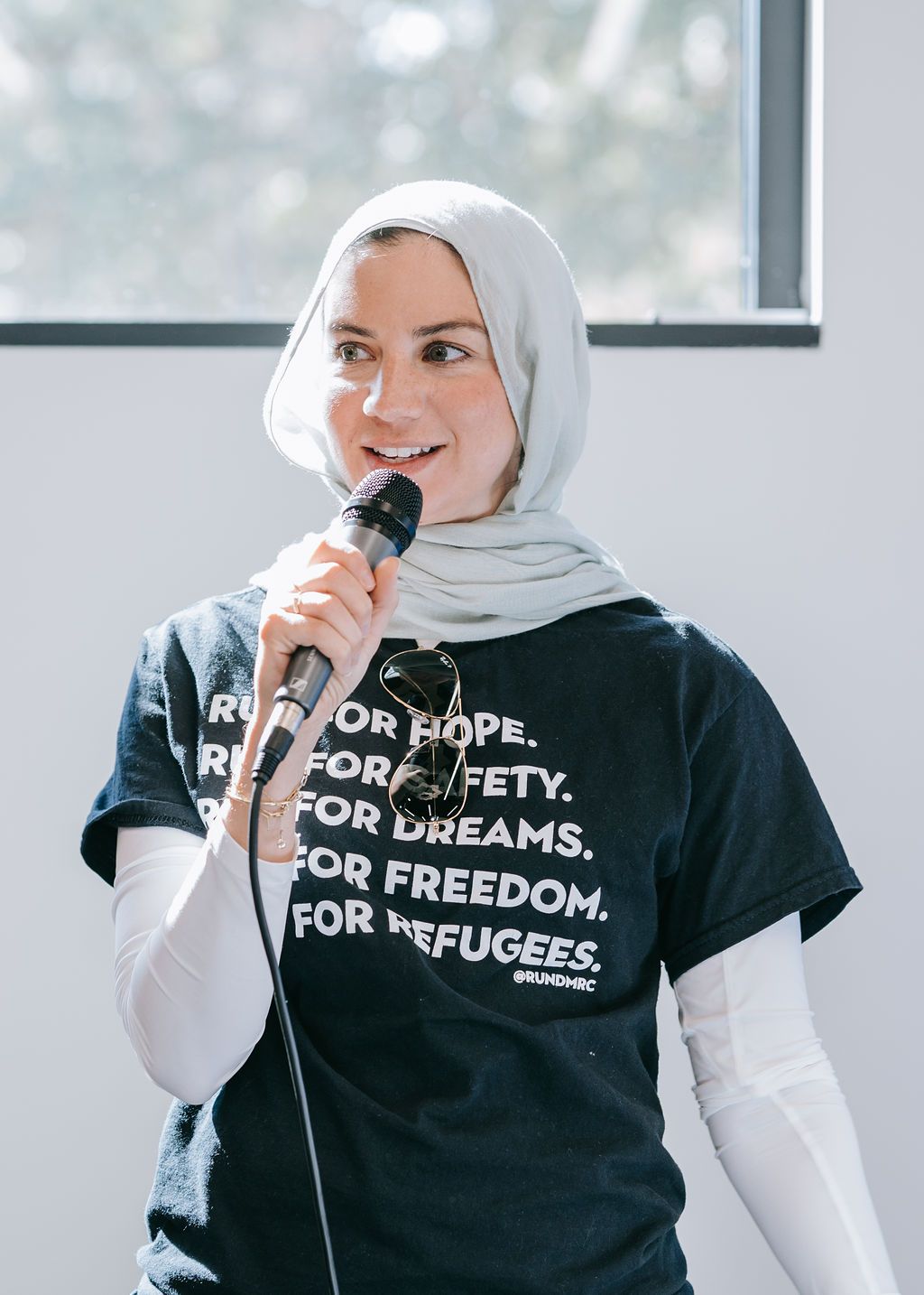 Person in hijab speaking into microphone wearing t-shirt with text about hope and refugees.