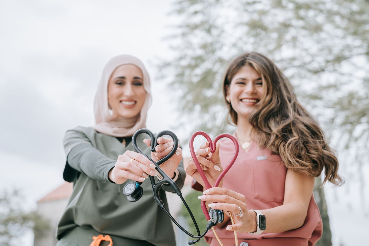 Two people making heart shapes with their hands against a cloudy sky outdoors.
