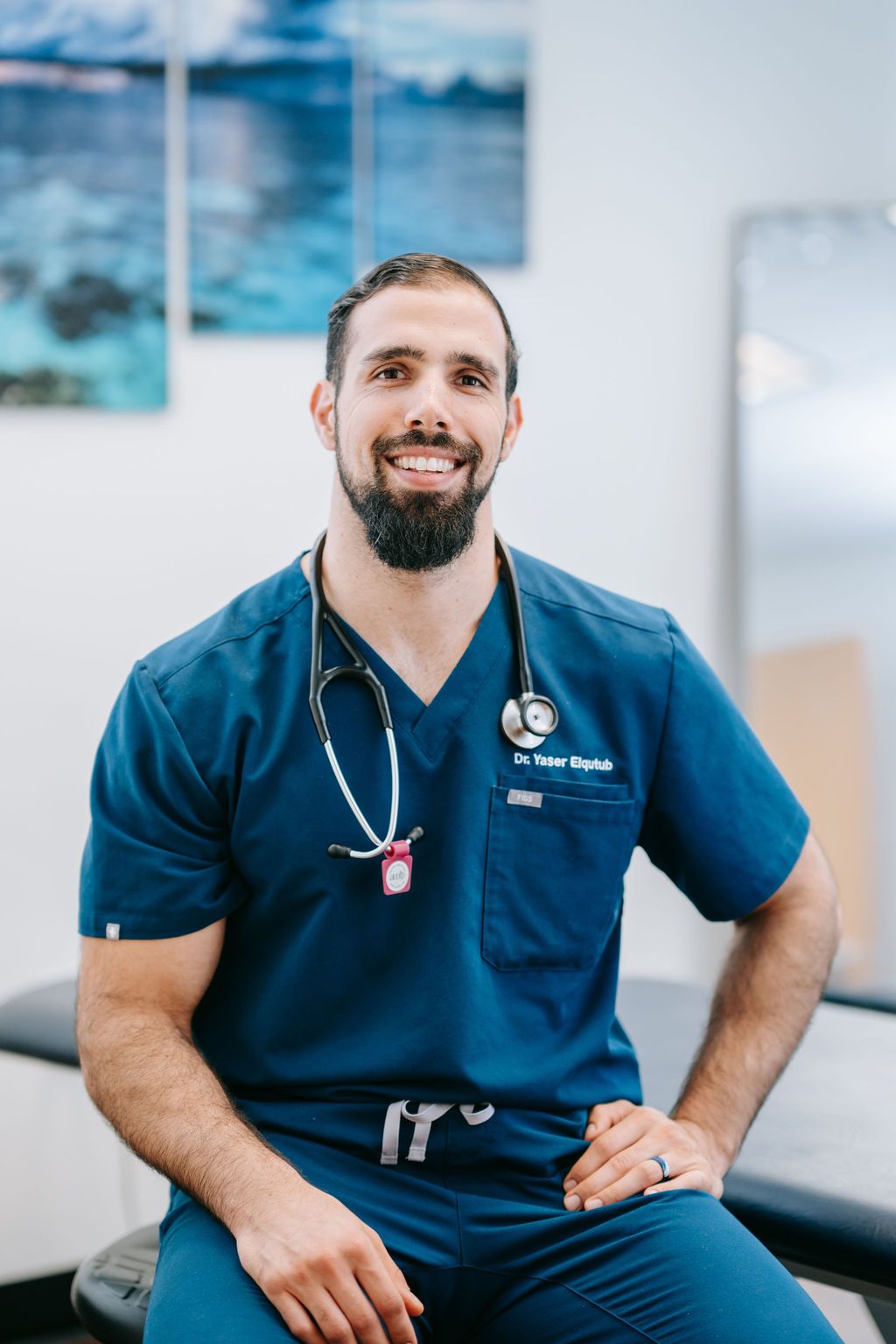 Medical professional in navy blue scrubs and stethoscope sitting in examination room with ocean artwork.