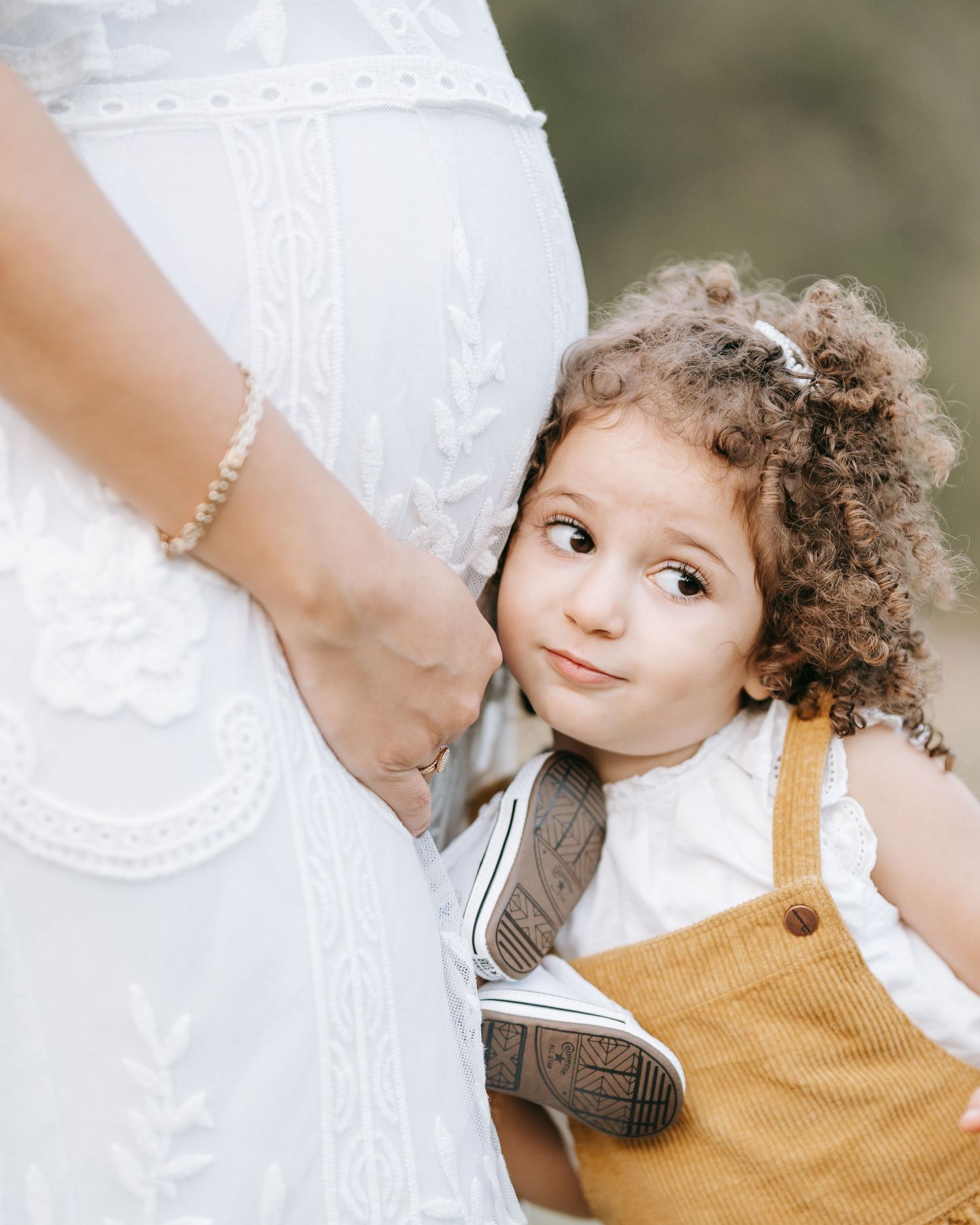 A child in yellow corduroy overalls stands next to someone wearing a white embroidered dress.