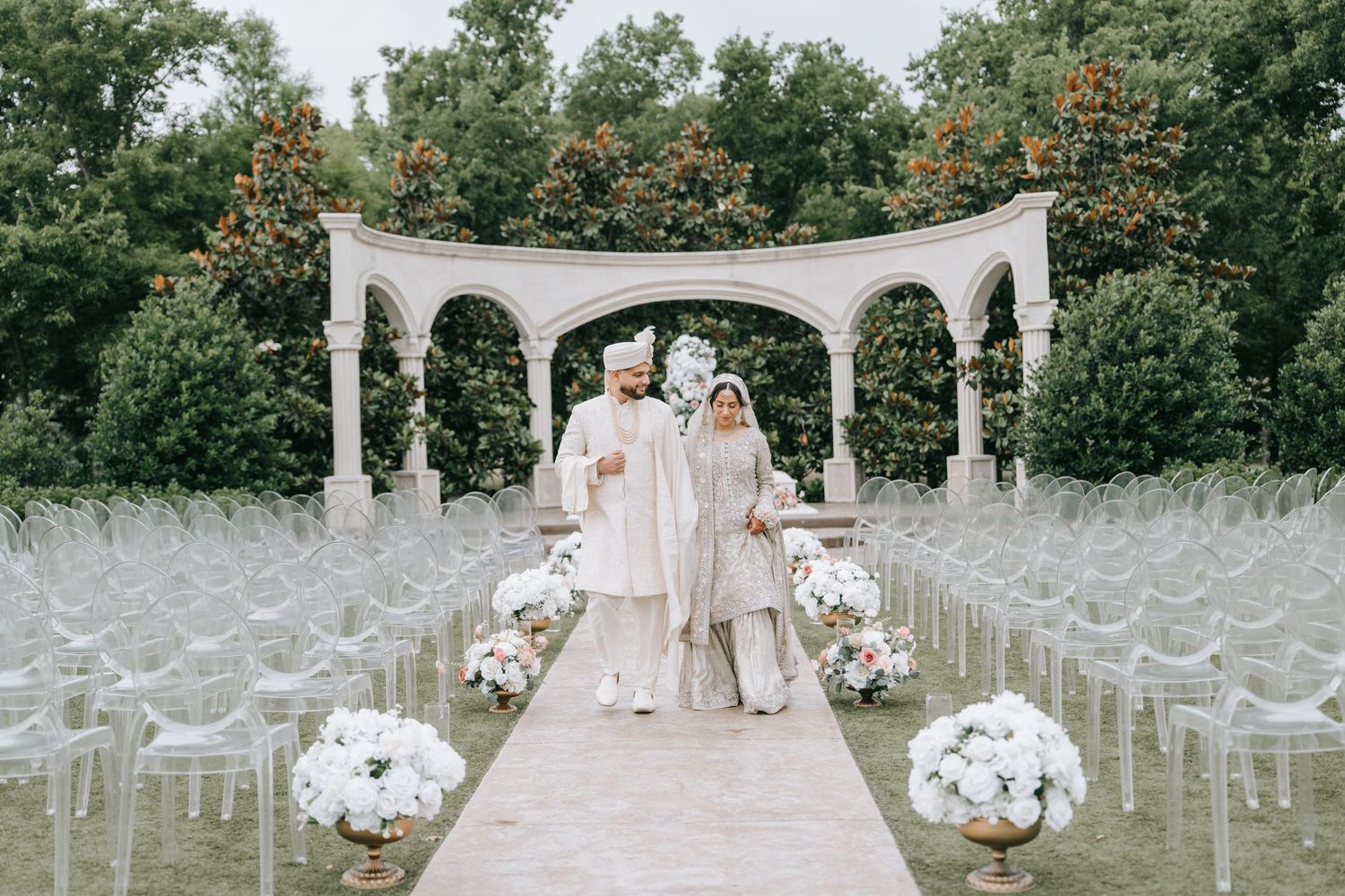 Elegant outdoor wedding ceremony setup with white floral arrangements and arched gazebo on stone pathway.