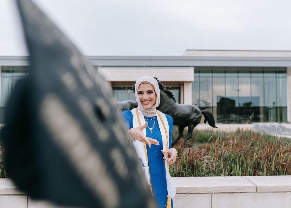 Graduate in academic regalia poses with black dog outside modern building.