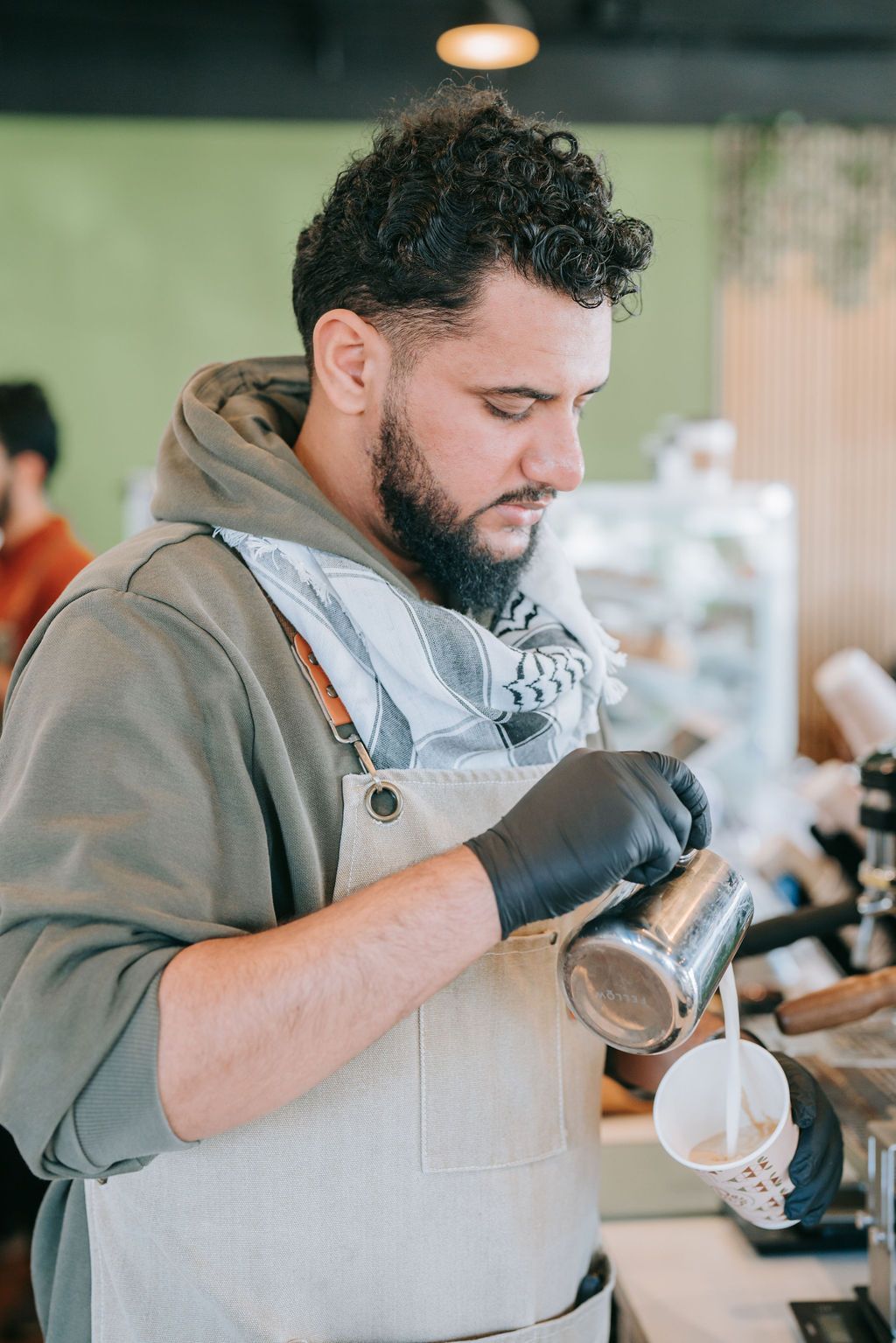 Barista in grey apron and hoodie steams milk at coffee station.