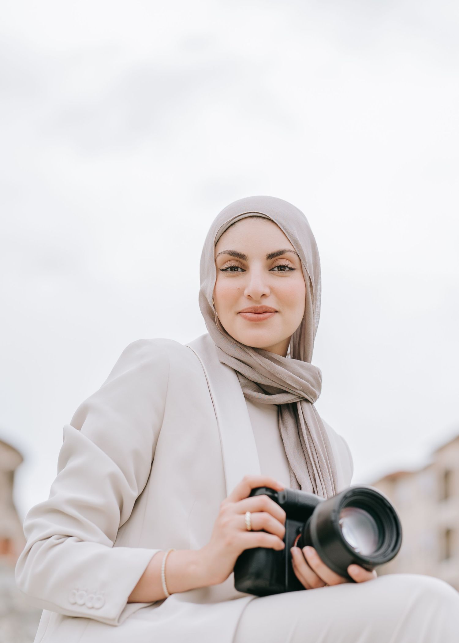 Professional photographer wearing neutral toned outfit and hijab holding DSLR camera outdoors.