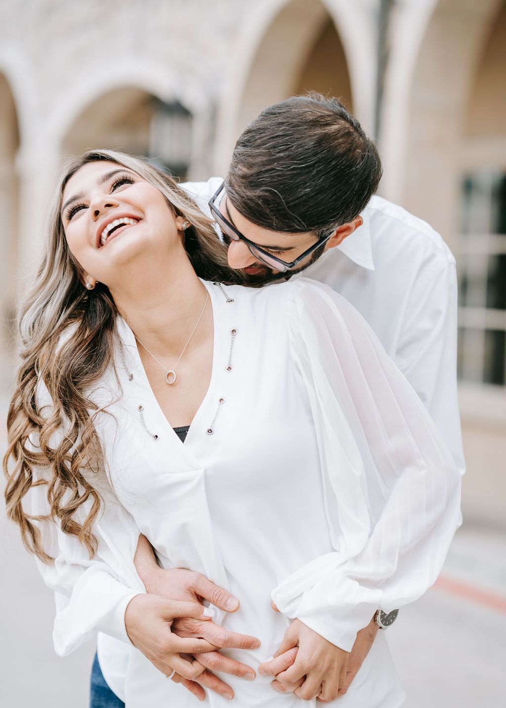 Romantic embrace between couple in white clothing against arched colonnade background.