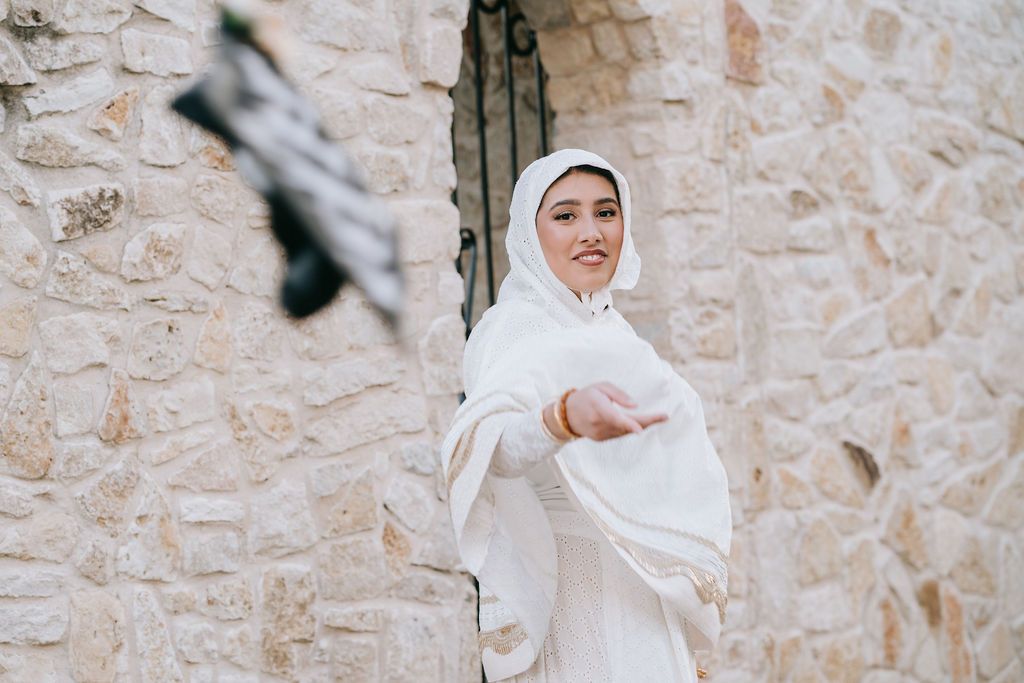 Graduate in traditional white dress poses elegantly against stone wall architecture.