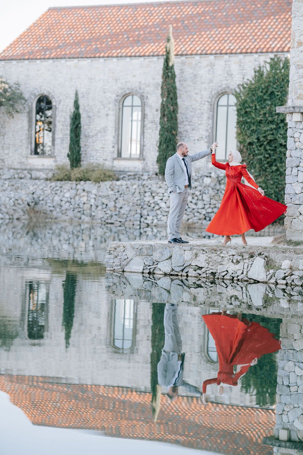 Romantic dance pose reflected in water by historic stone building with cypress trees.