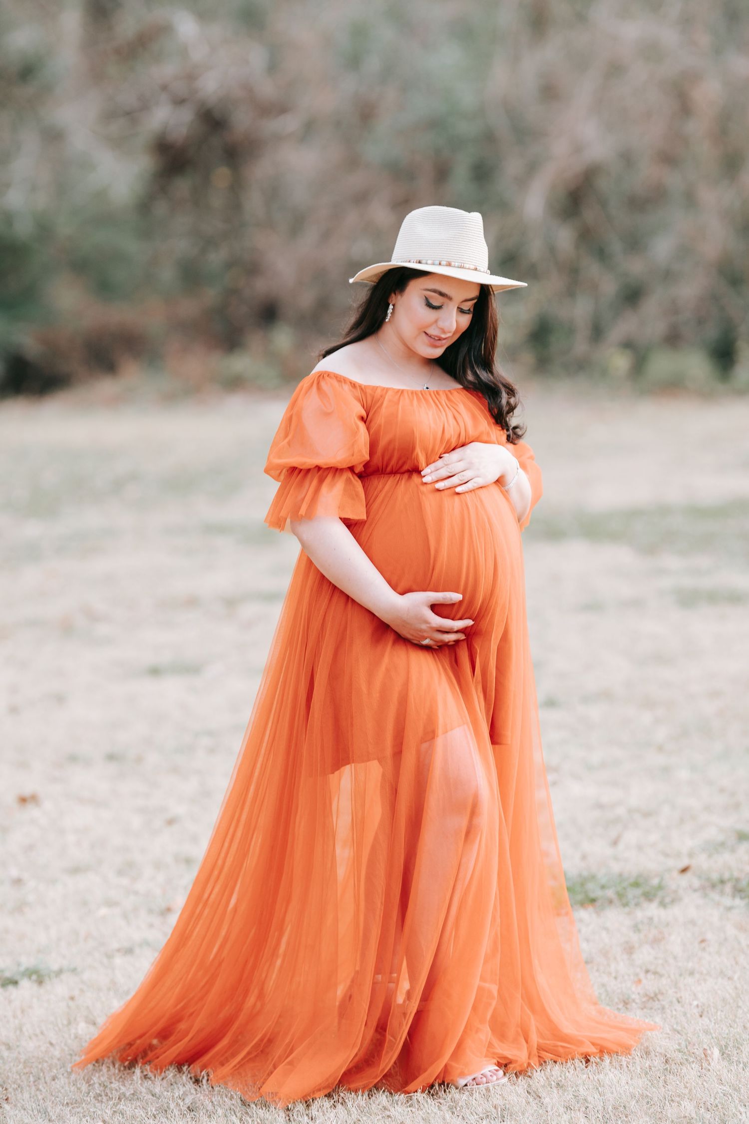 A pregnant woman in an orange gown and hat poses gracefully in a natural setting.