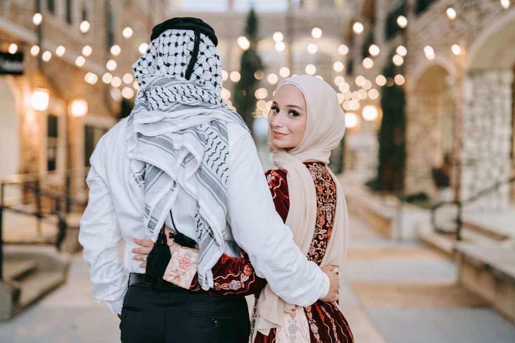 A couple in traditional Middle Eastern dress posing together under string lights in an elegant corridor.
