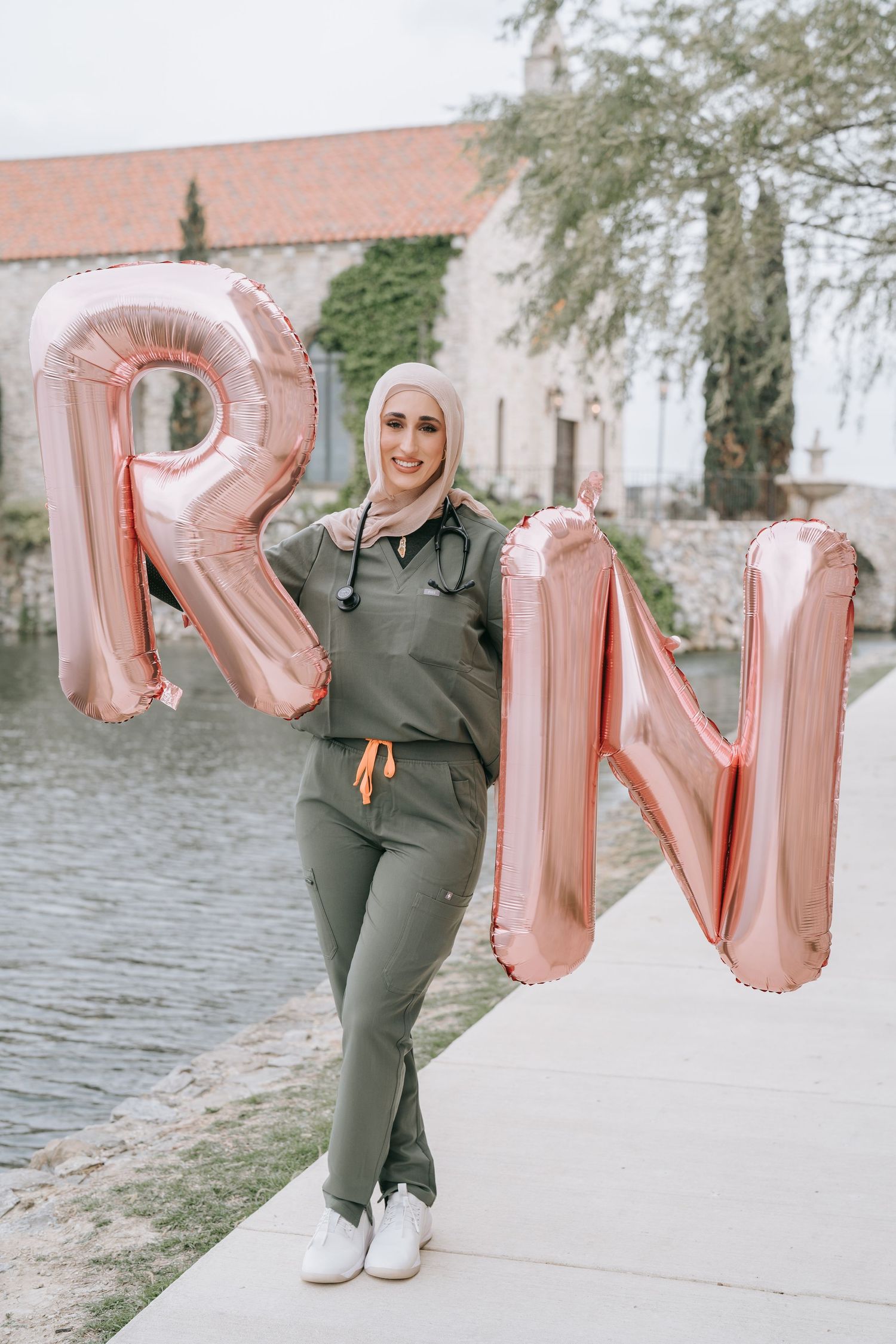 Person holding rose gold letter balloons spelling 'RN' outdoors near a stone building with ivy.