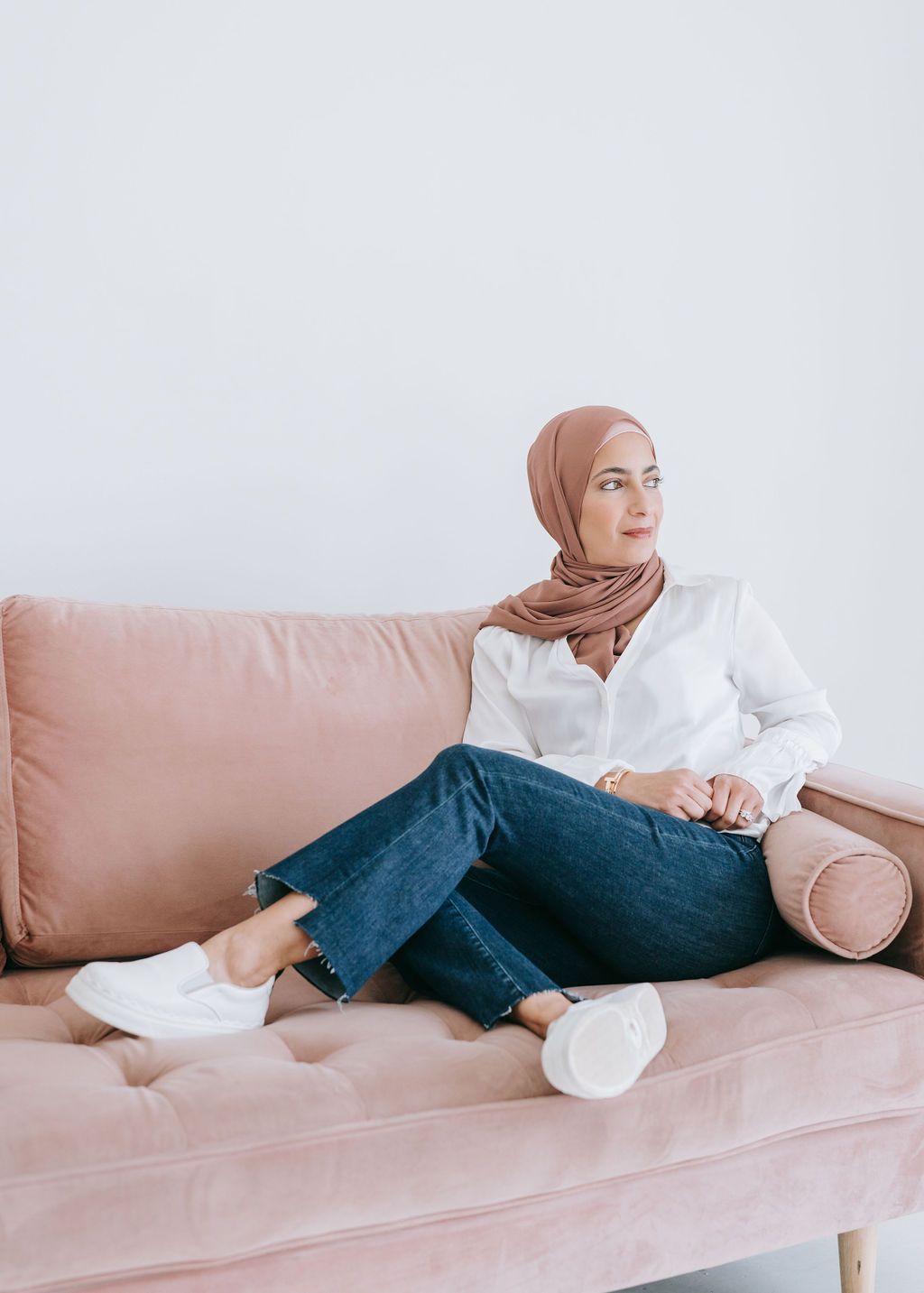 Woman in hijab and casual denim relaxing on pink couch in minimalist setting.