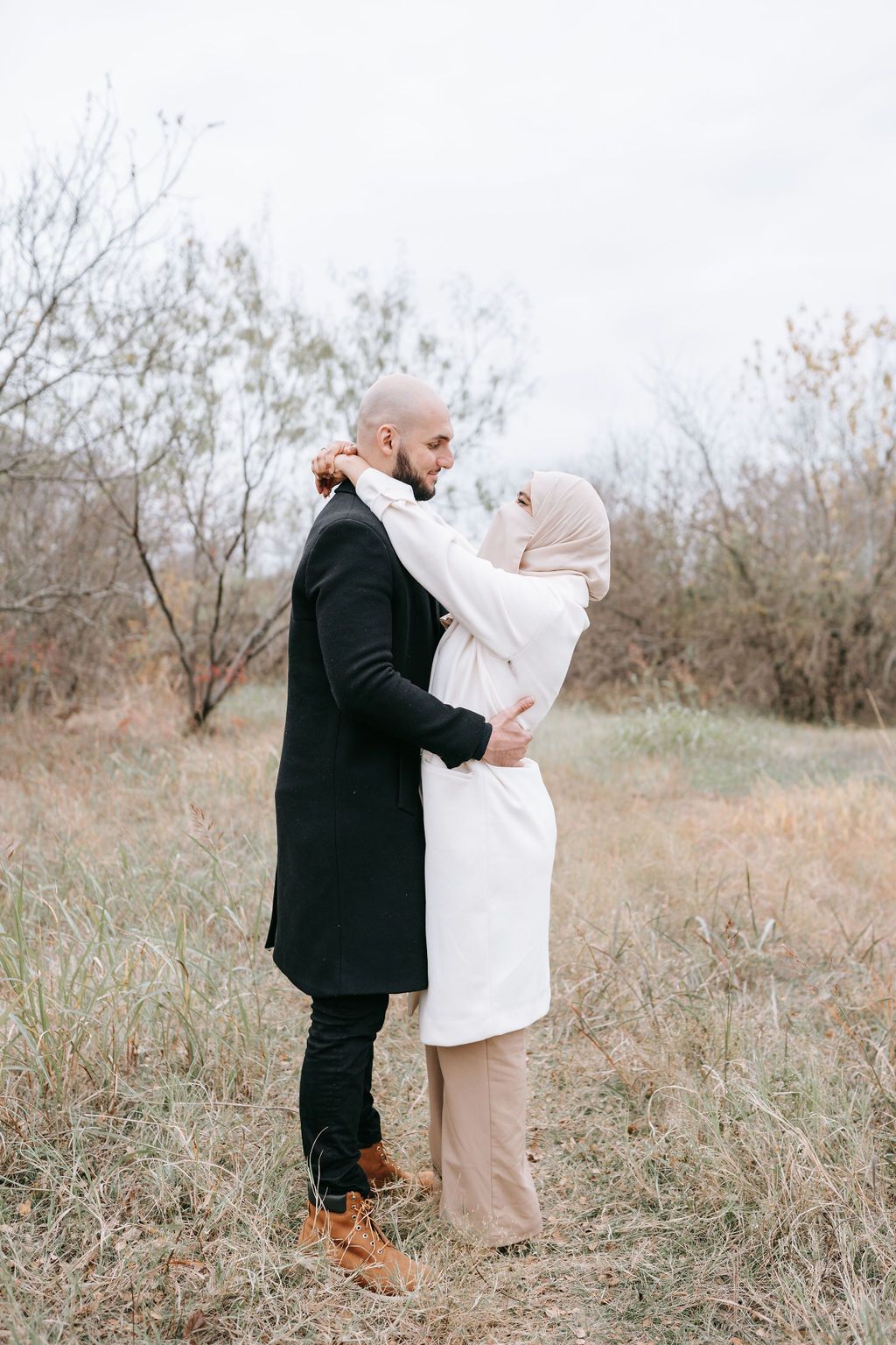 Winter portrait of couple embracing in casual attire against bare branches.