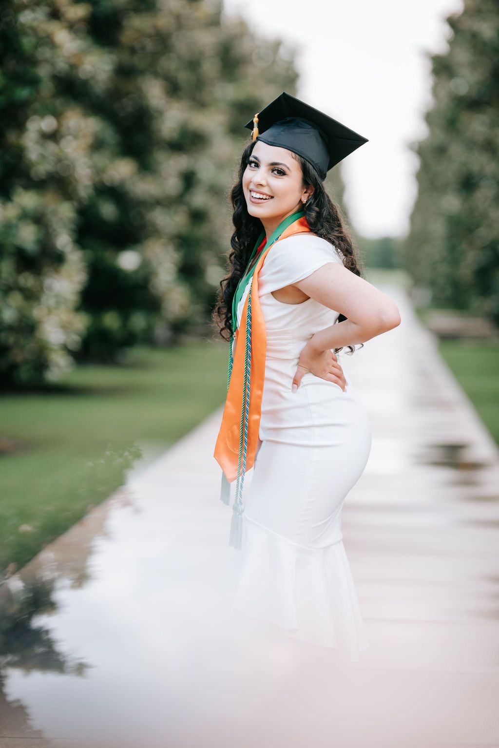 Graduate in cap and gown with tassel poses on walkway during outdoor photoshoot.