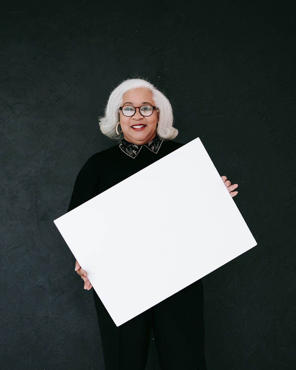 Person in black outfit holding blank white sign board against dark background.