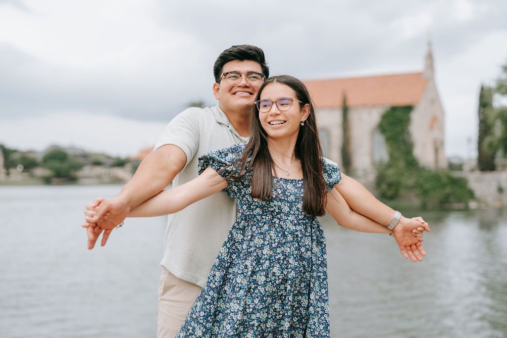 A carefree couple poses with outstretched arms by a lakeside with a building in the background.