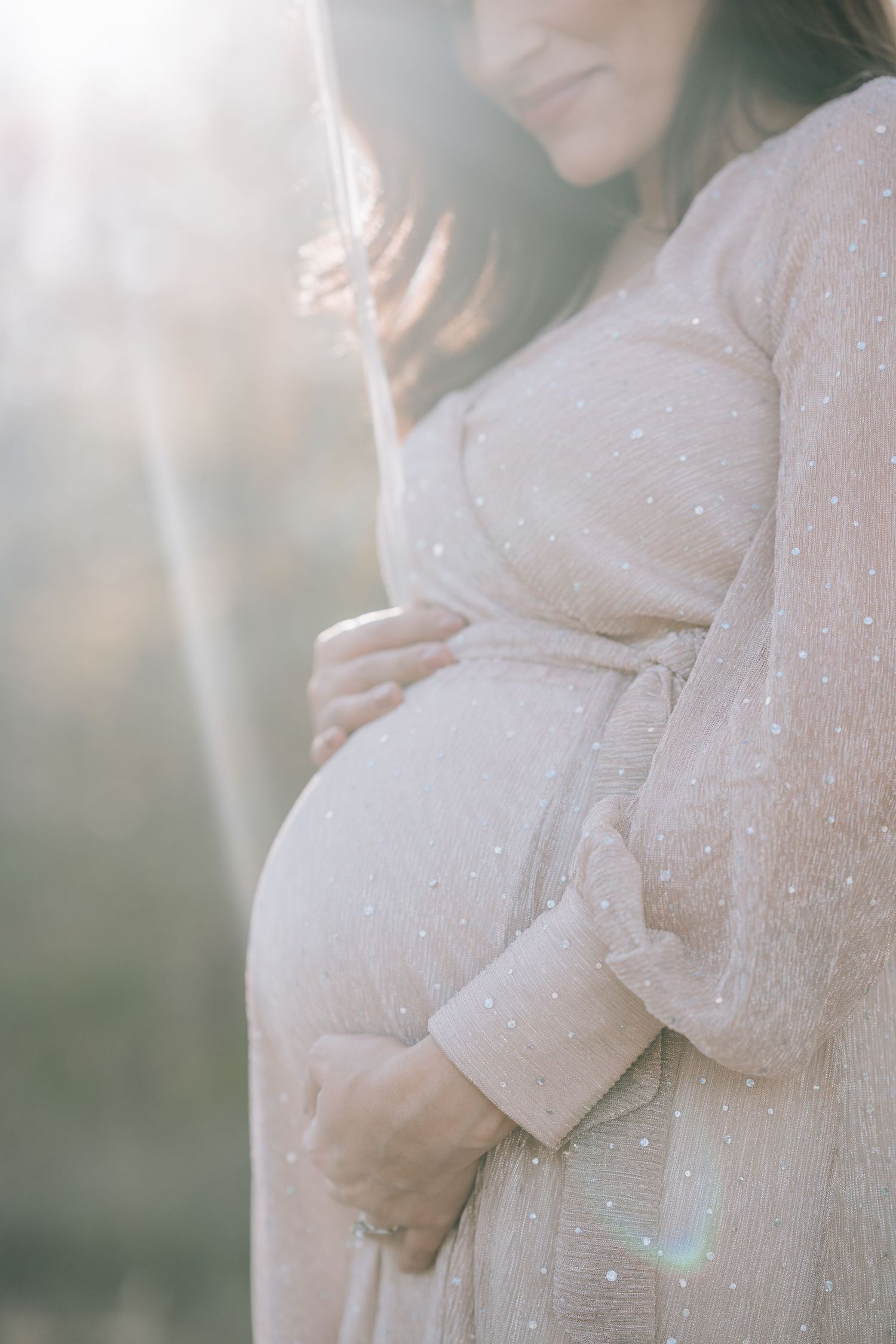 A close-up shot of a pregnant belly in a sparkly blush dress with sunlight creating a dreamy effect.