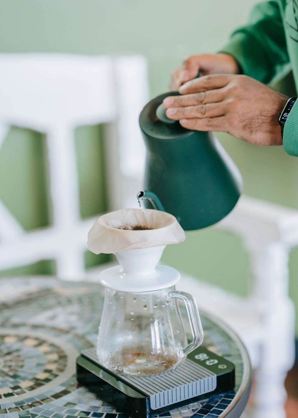 Person pouring hot water over coffee filter in pour-over setup with green background.