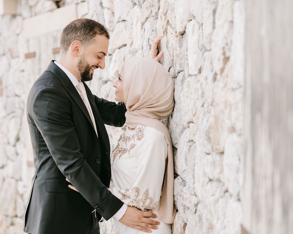Elegant couple shares intimate moment by stone wall with bride in hijab.