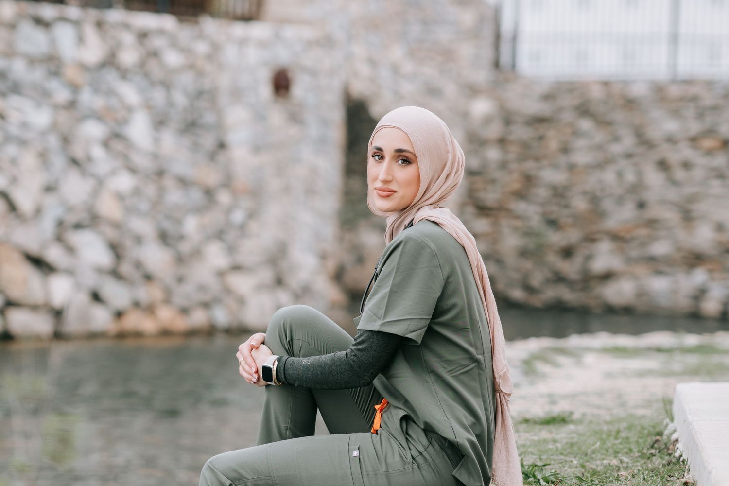Woman in green outfit sits thoughtfully by stone wall near water.