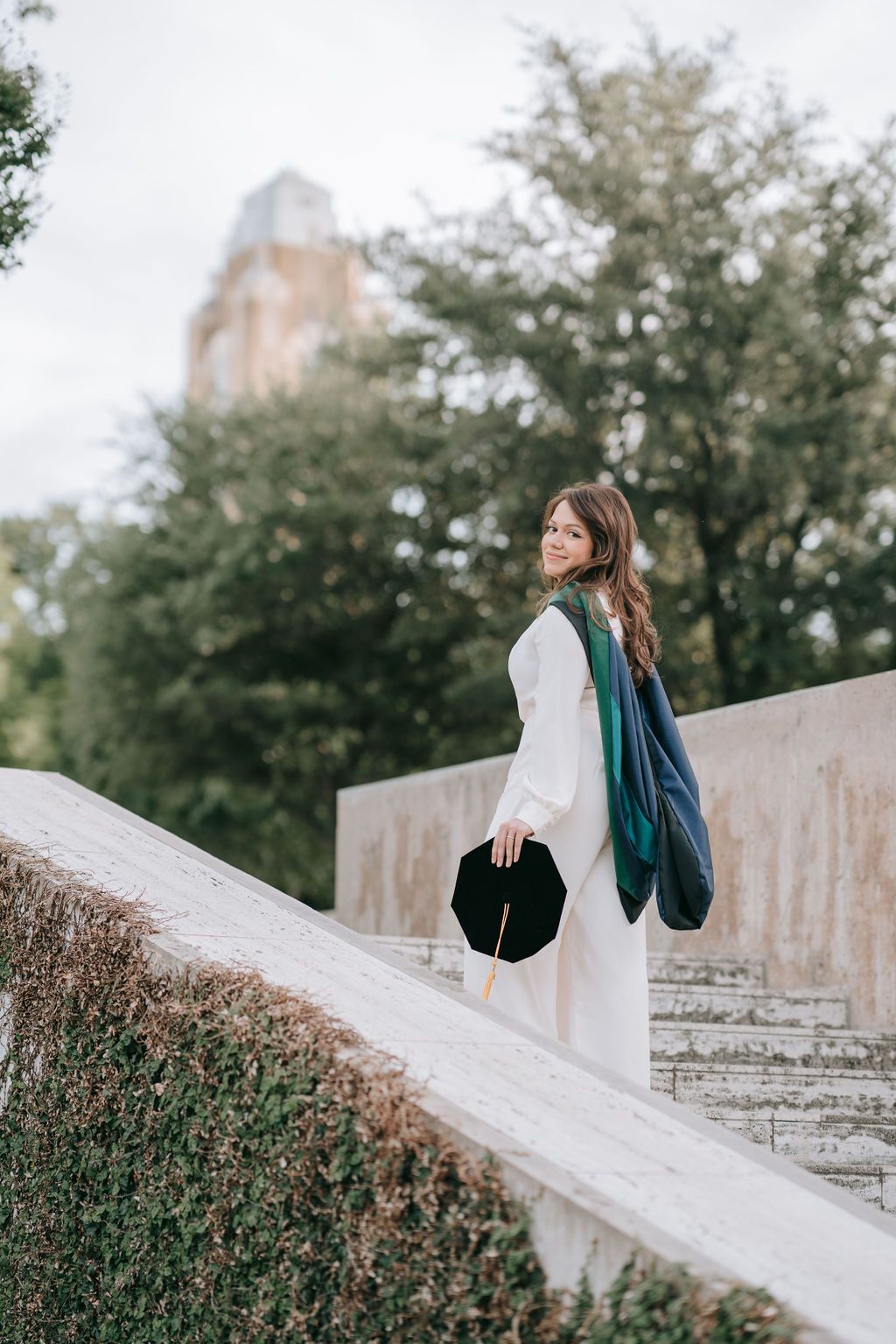 A graduate in white posing on concrete stairs with a historic building tower in background.