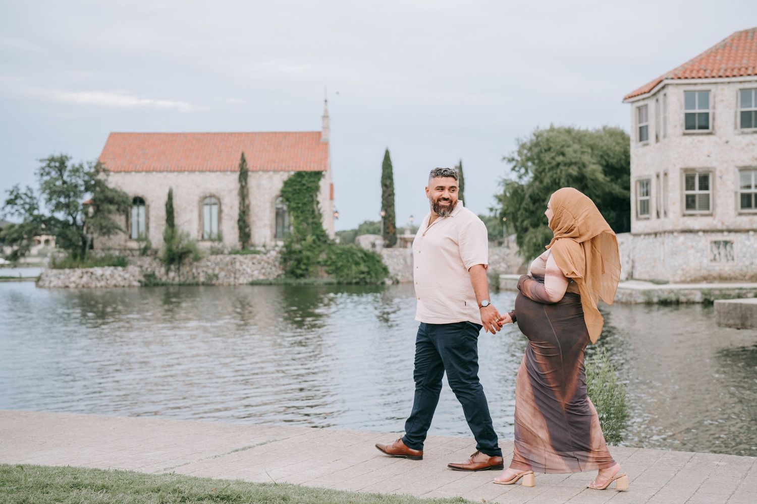 A couple strolls along a waterfront path with Mediterranean-style buildings in the background.