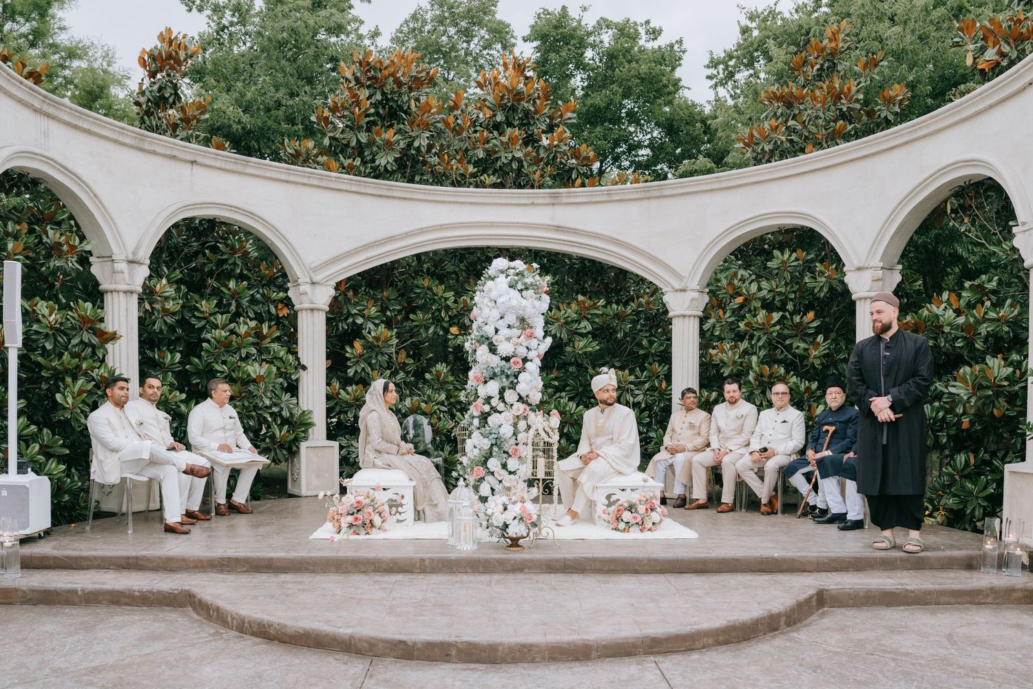 Wedding party wearing light colored outfits poses in front of green trees with orange flowers in outdoor arched venue.
