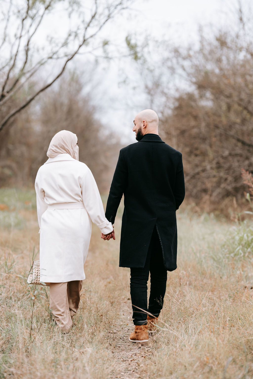 Couple holding hands while walking through autumn field in coats.