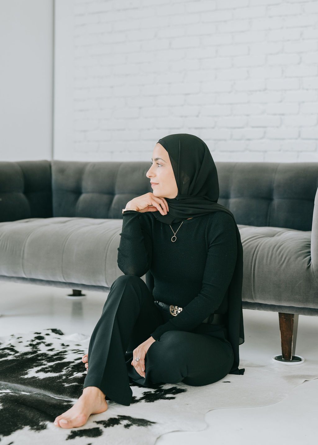 Person in black hijab and outfit sits relaxed on grey couch against white brick wall.