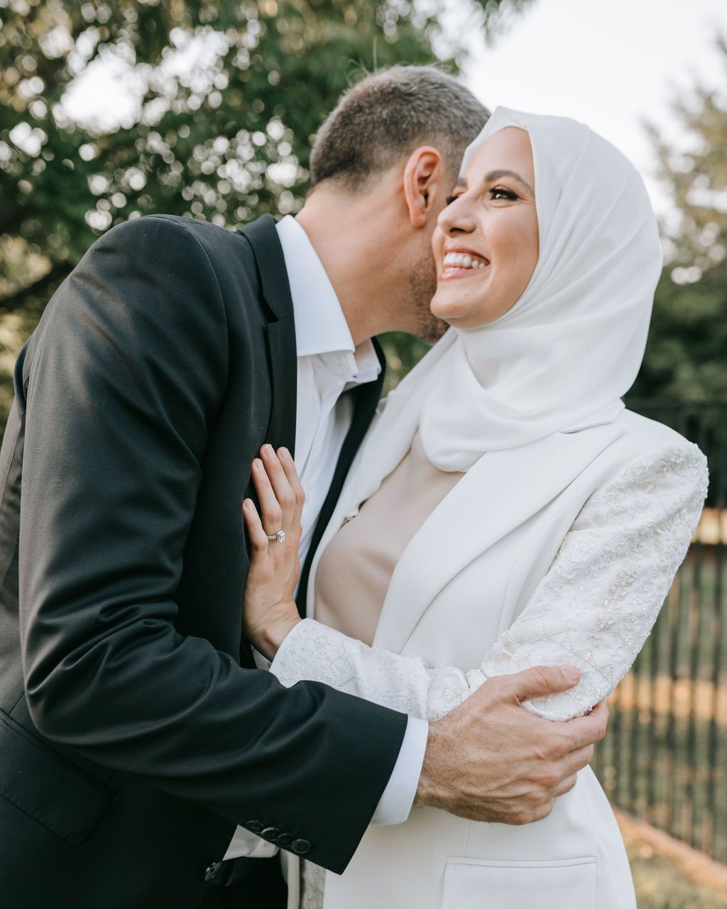 A romantic embrace between newlyweds in formal attire against a green outdoor background.