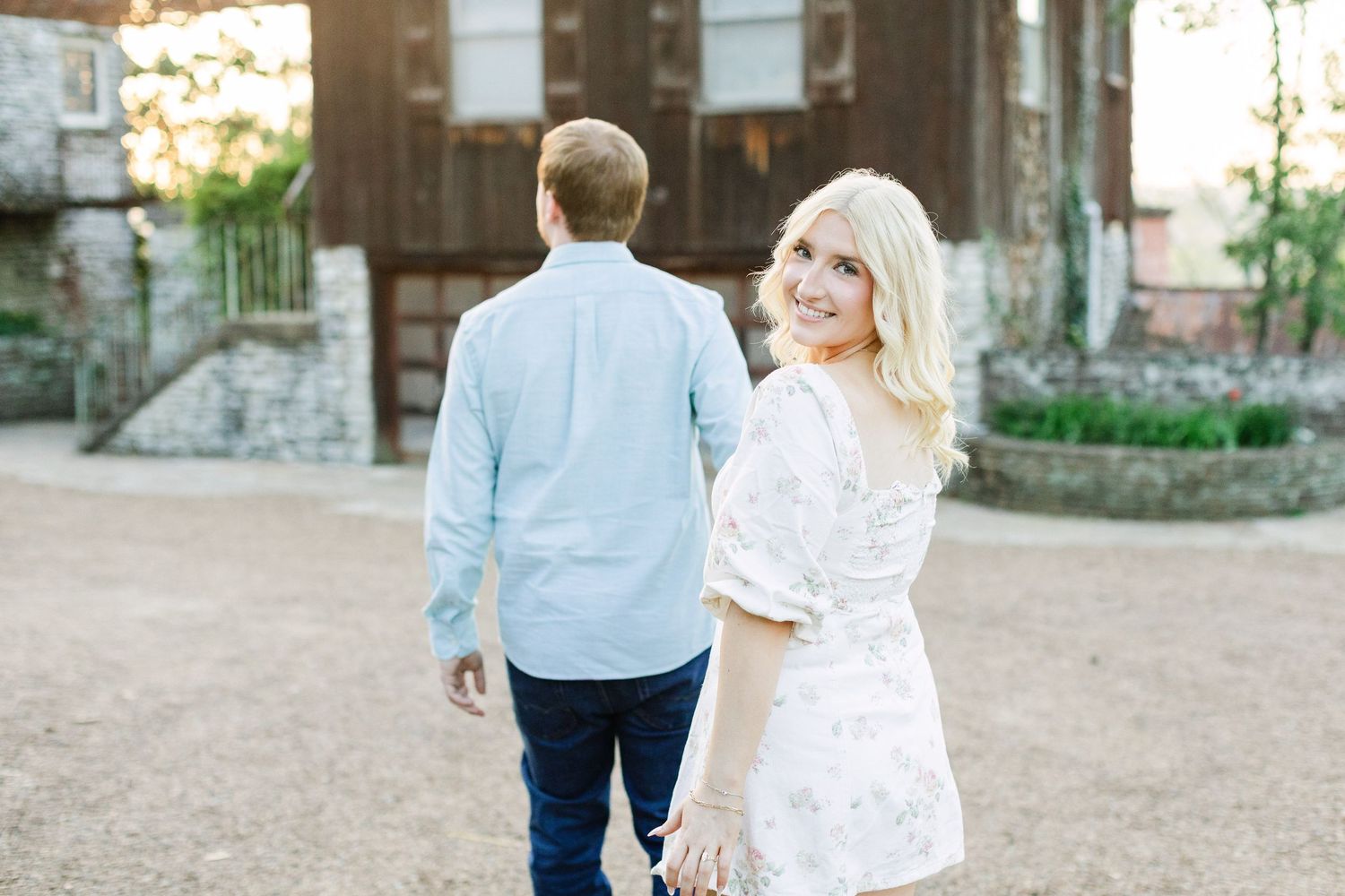 Blonde woman in white floral dress smiles over shoulder while holding hands with man in blue shirt outdoors.