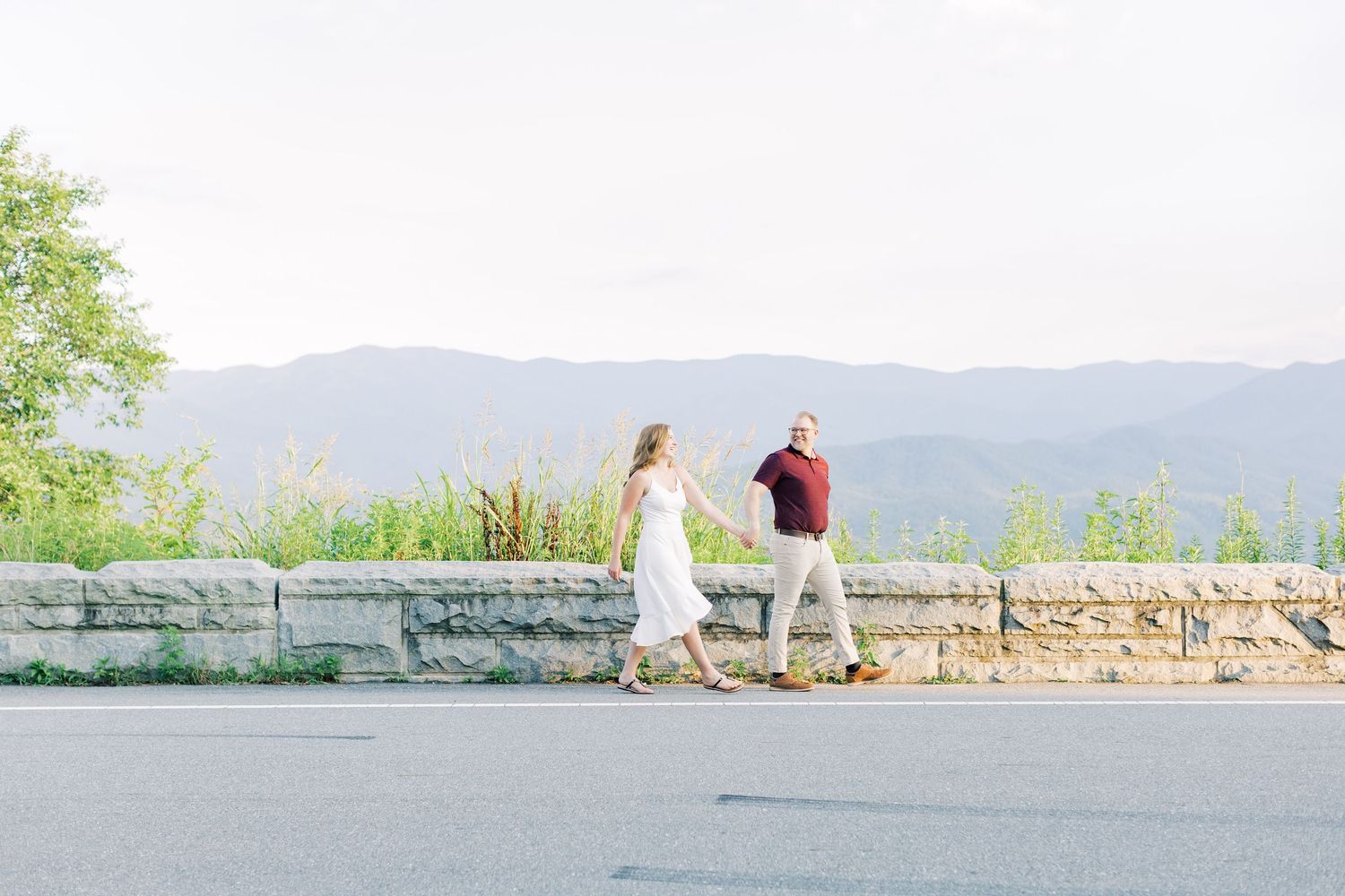 Couple walking hand in hand along a stone wall overlook with scenic mountain views in the background.