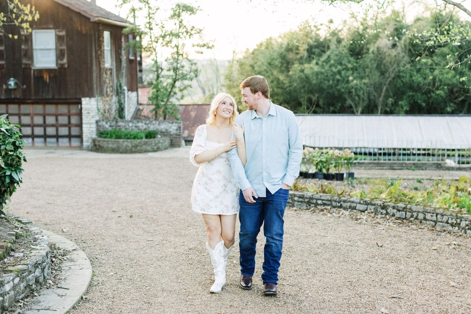 A smiling couple walks hand-in-hand on a gravel path near a rustic barn, woman in white dress and boots.