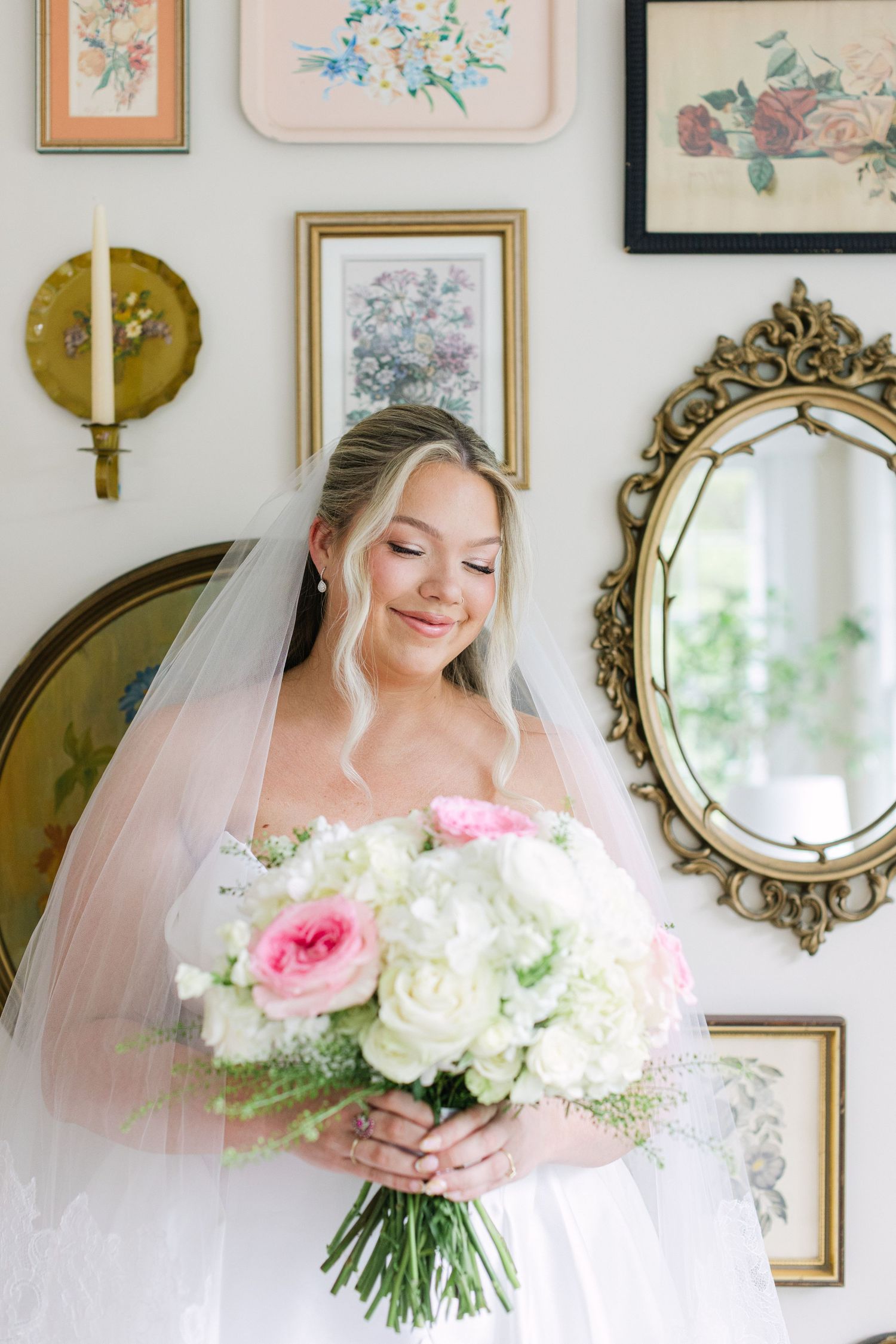 Bride in white gown and veil holds pink and white rose bouquet, smiling softly in a vintage-styled room.