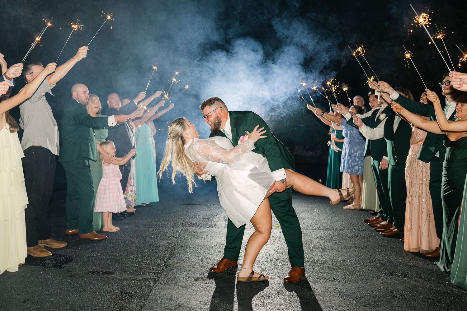 Bride and groom share romantic dip kiss during sparkler exit at night wedding reception with guests cheering.