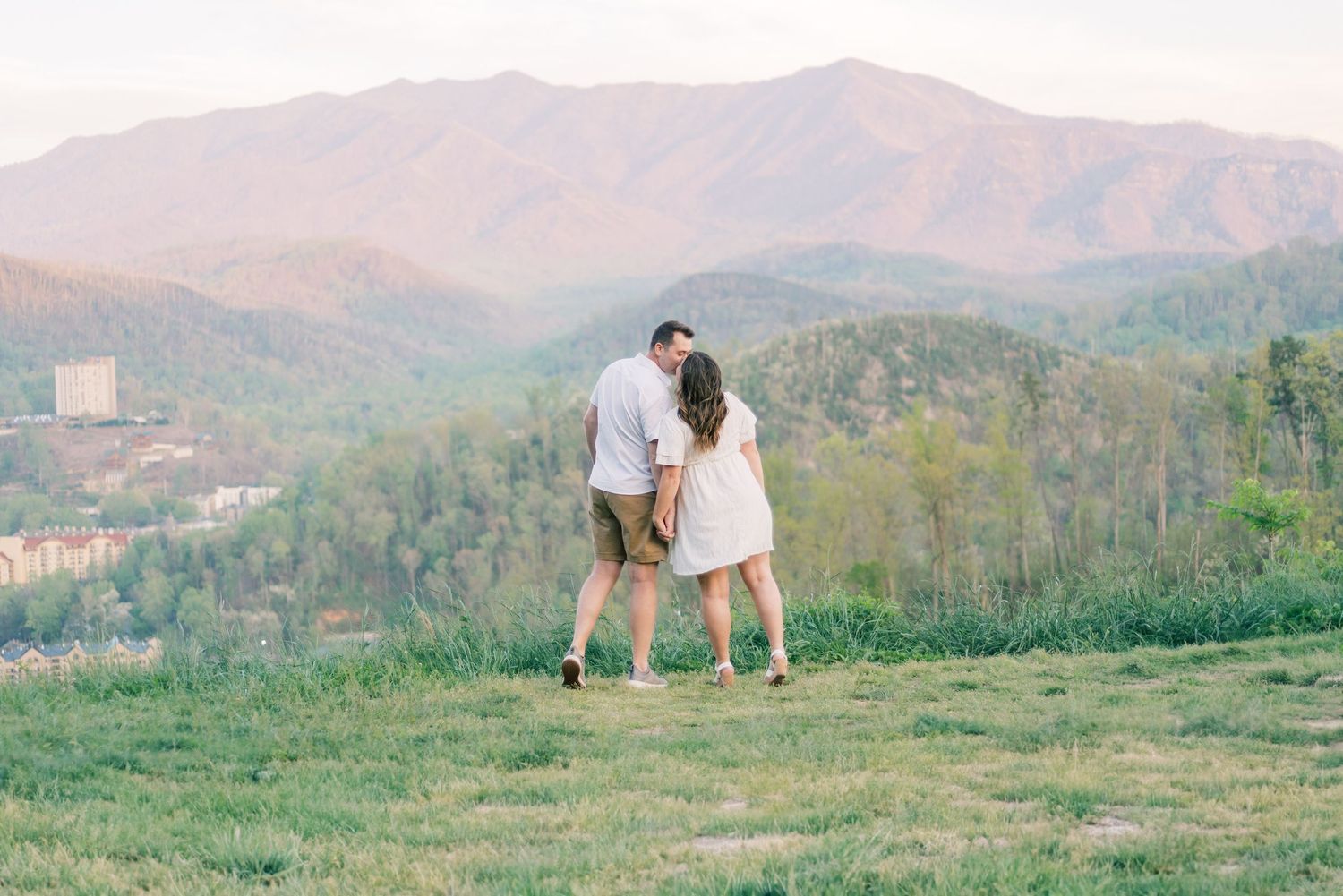 Couple holding hands on a grassy hilltop with stunning mountain views and soft sunset light in the background.