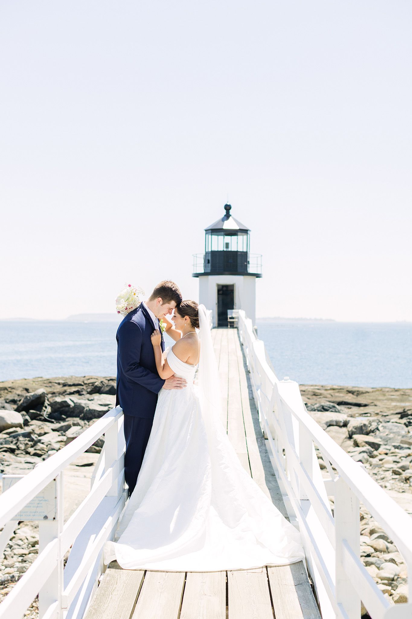 Bride and groom share a romantic moment on a white boardwalk leading to a coastal lighthouse on a sunny day.