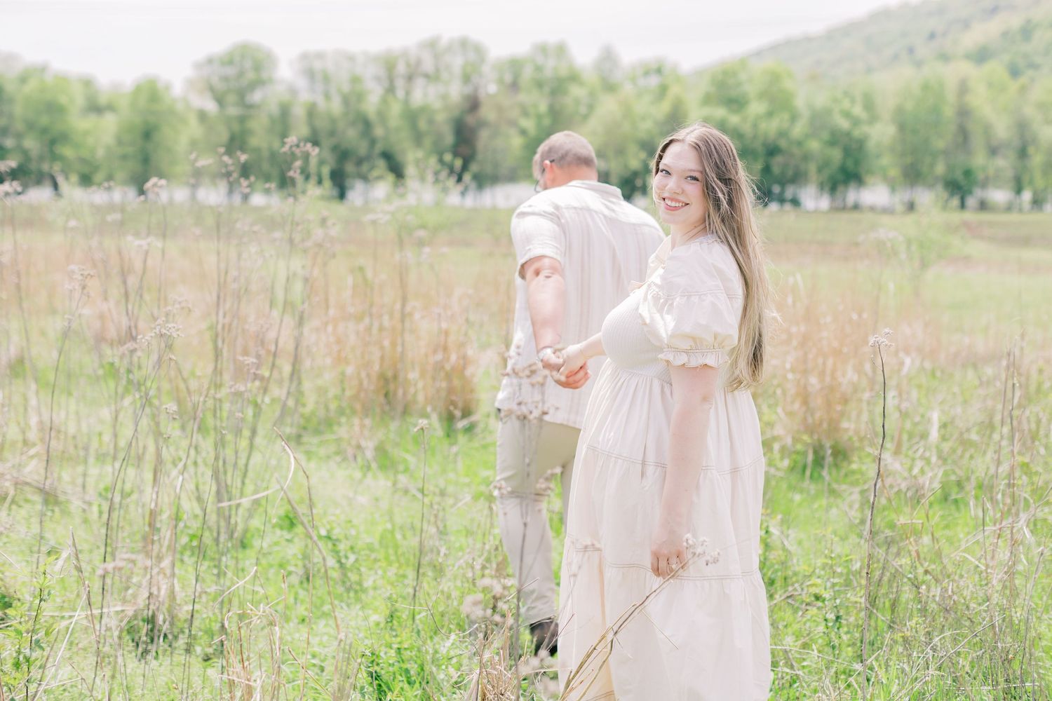 Couple in white outfits standing in a lush green meadow, woman smiling over shoulder, mountains in background.