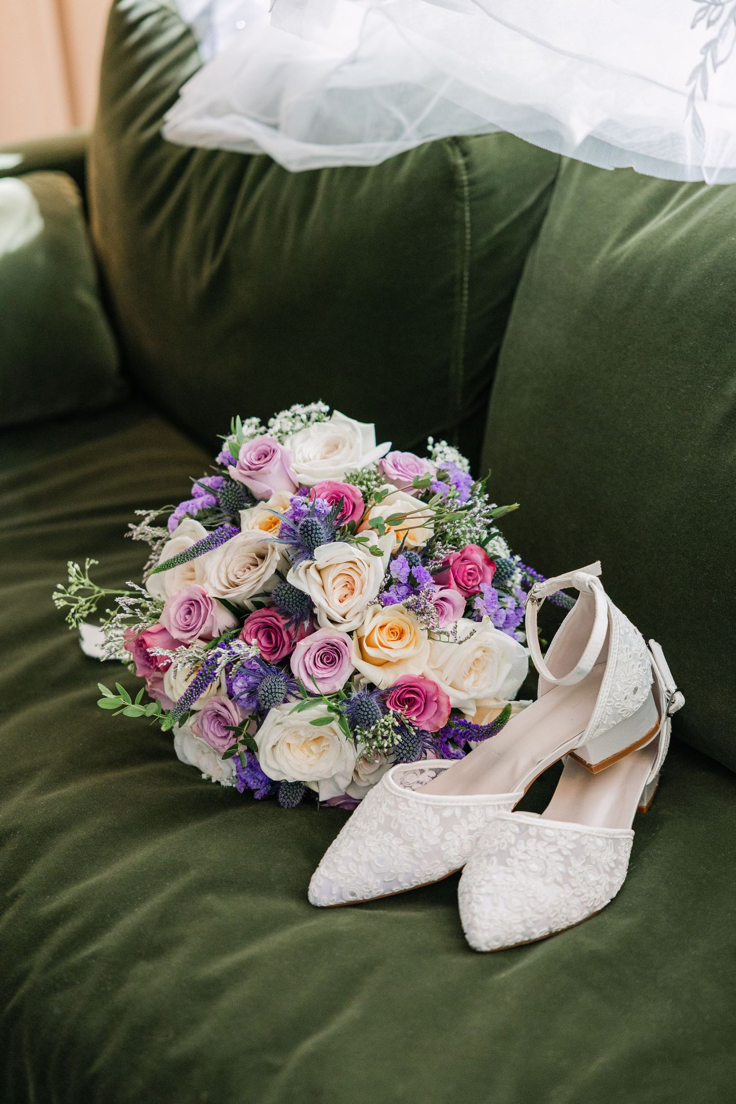 Bridal bouquet of pink, purple, and cream roses beside white lace heels on a green velvet sofa.