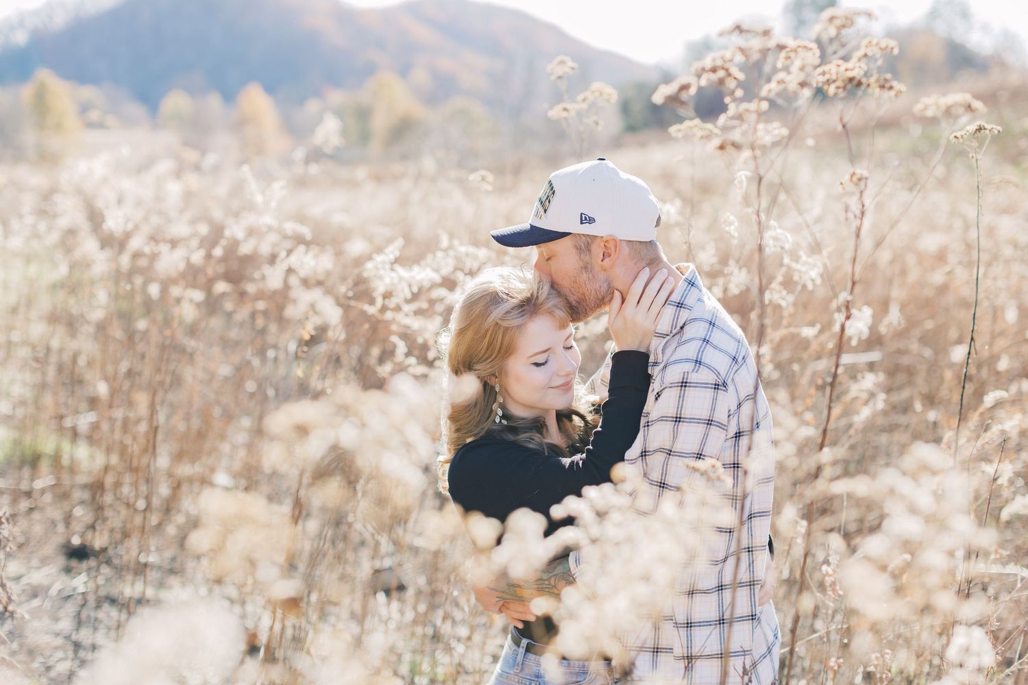 Couple embracing romantically in a golden field of tall grasses with mountains in the background during autumn.