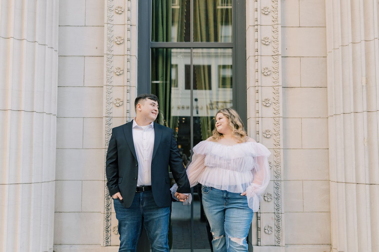 Couple holding hands and smiling at each other in front of elegant stone columns during an outdoor engagement photo session.